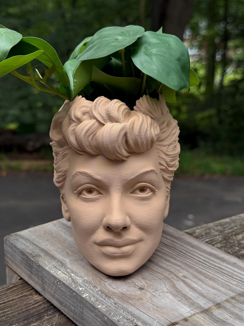 May include: A beige-colored planter shaped like a woman's head, with detailed facial features and curly hair. The top of the head is open, holding green leafy plants. The planter is sitting on a wooden surface.