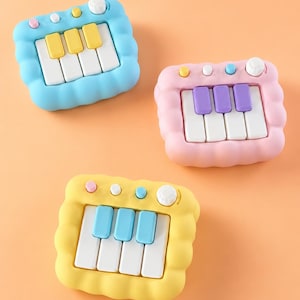 May include: Three colorful toy pianos in blue, pink, and yellow. Each has a white and colored key layout, with a knob and buttons on top. The blue piano has yellow keys, the pink has purple, and the yellow has blue.