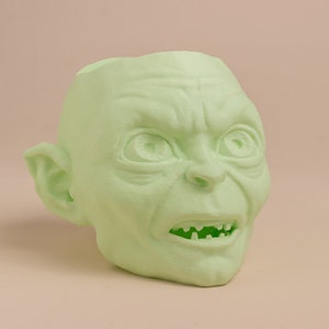 May include: A green, 3D printed planter shaped like the head of Gollum from the Lord of the Rings. The planter has a wide mouth and a detailed face with a frowning expression.