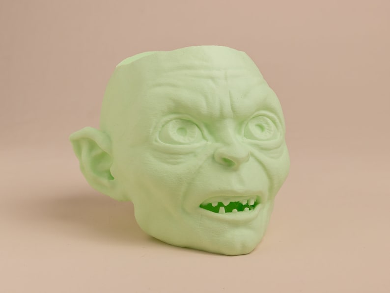 May include: A green, 3D printed planter shaped like the head of Gollum from the Lord of the Rings. The planter has a wide mouth and a detailed face with a frowning expression.