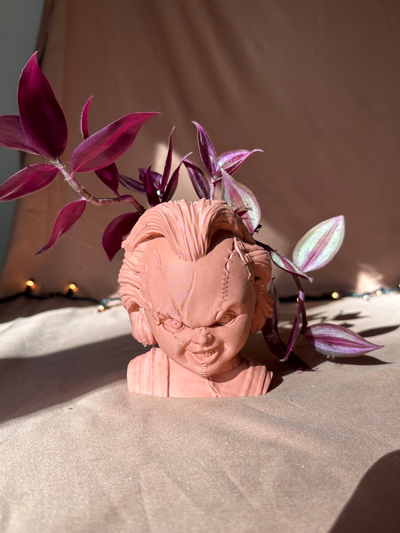 May include: A terracotta-coloured planter shaped like the head of a horror movie character with a stitched-up face. The planter is filled with a leafy green plant with purple leaves.