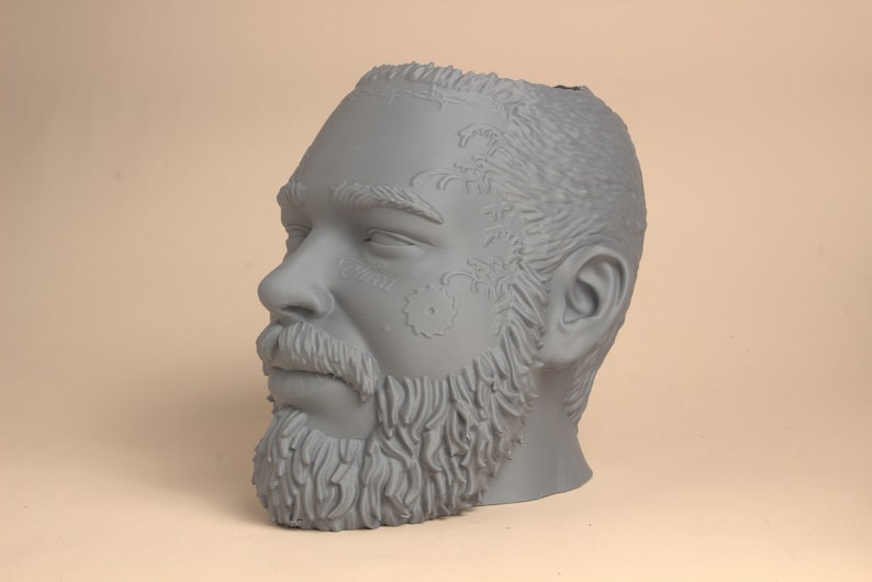 May include: A gray 3D printed sculpture of a man's head with a beard and tattoos. The head is tilted to the side and the man's eyes are closed.