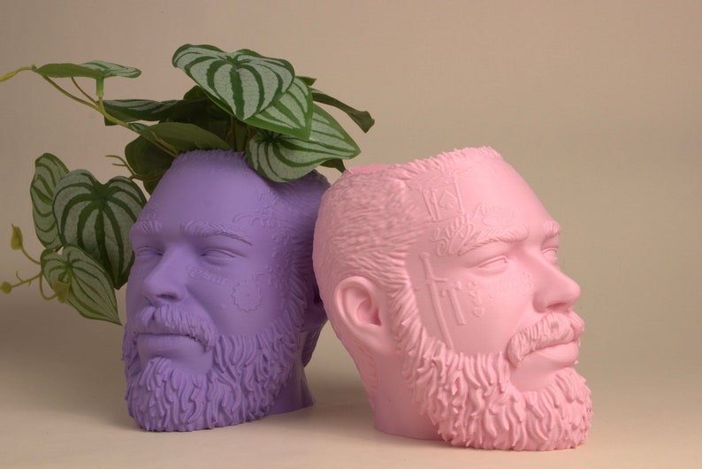 May include: Two ceramic planters shaped like human heads, one is purple and the other is pink. Both planters have detailed facial features and beards. The purple planter has a green plant growing out of the top.