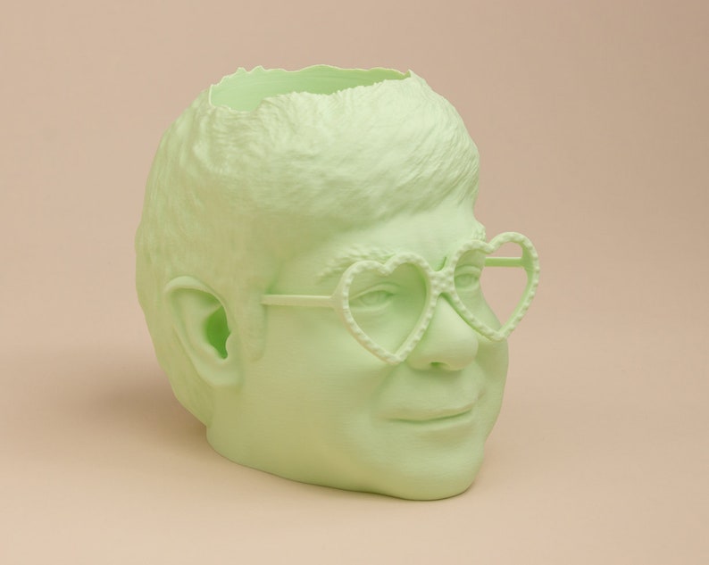 May include: A light green ceramic planter shaped like a person's head with a broken top, wearing heart-shaped glasses.