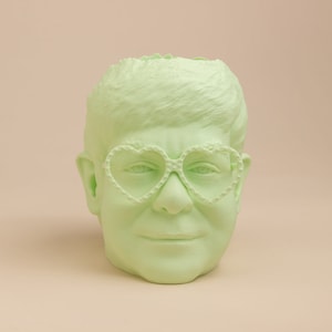 May include: A light green ceramic planter shaped like a person's head with heart-shaped glasses.