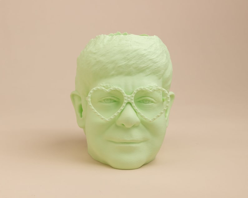 May include: A light green ceramic planter shaped like a person's head with heart-shaped glasses.