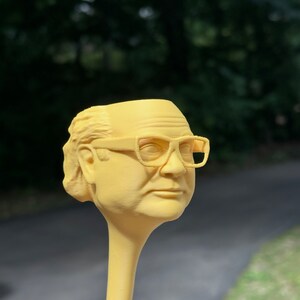 May include: A yellow plastic figurine of a person wearing glasses. The figurine is attached to a long, yellow, plastic stick.