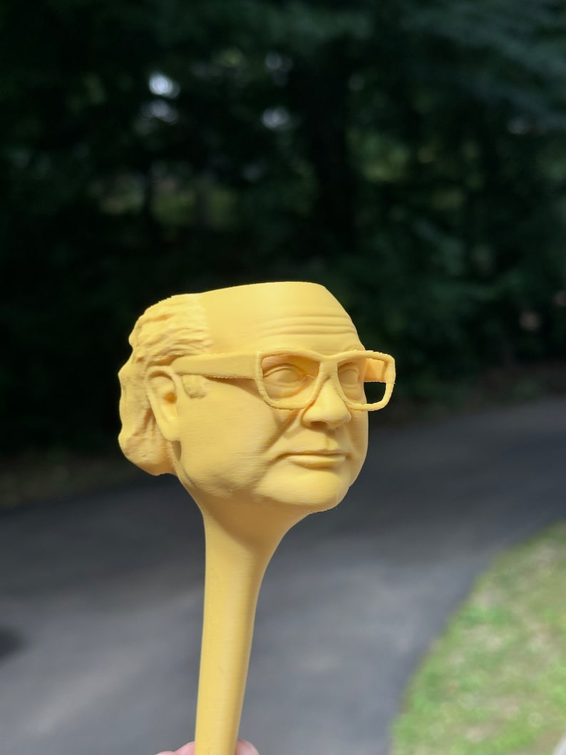 May include: A yellow plastic figurine of a person wearing glasses. The figurine is attached to a long, yellow, plastic stick.