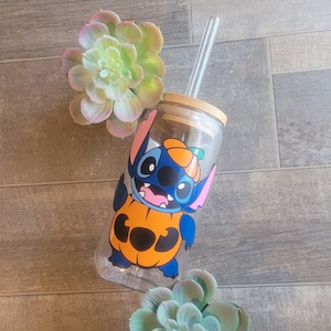 May include: A clear glass tumbler with a blue and orange cartoon character wearing a pumpkin costume. The character has a stitched design and the text "BOO" is on the pumpkin. The tumbler has a straw and a wooden lid.