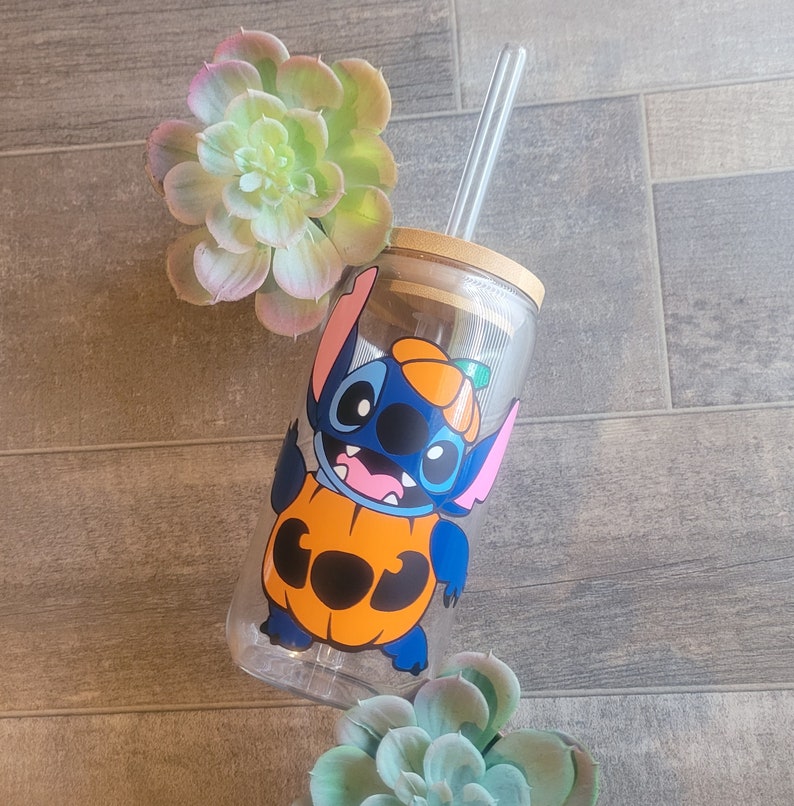 May include: A clear glass tumbler with a blue and orange cartoon character wearing a pumpkin costume. The character has a stitched design and the text "BOO" is on the pumpkin. The tumbler has a straw and a wooden lid.