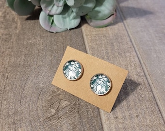 Może przedstawiać: Para kolczyków z zielonym i białym logo Starbucks na każdym kolczyku. Kolczyki znajdują się na brązowej kartce.