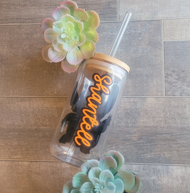 May include: A clear plastic tumbler with a black silhouette of a ghost and the word "Haunted" in orange lettering. The tumbler has a wooden lid and a clear plastic straw.