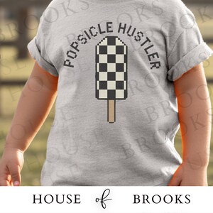 May include: A light gray t-shirt featuring the text "POPSICLE HUSTLER" arched above a pixelated popsicle design with a black and white checkered pattern. The popsicle is on a brown stick.