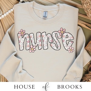 Peut inclure: Un sweat-shirt beige clair avec le mot "NURSE" imprimé en lettres noires soulignées. Les lettres sont remplies d'un motif de fleurs roses et de symboles médicaux.
