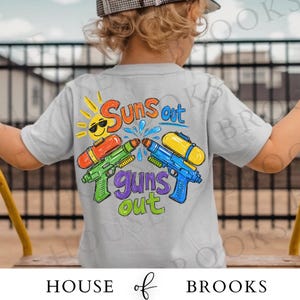 以下が含まれることがあります： 「Suns out guns out」と書かれたカラフルなグラフィックが特徴の、明るい灰色の子供用Tシャツ。 デザインは、笑顔の太陽、水鉄砲、水滴が特徴です。