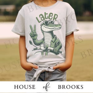 May include: A light gray t-shirt with a green alligator wearing sunglasses and riding a scooter. The alligator is giving a peace sign and the text "Later" is above the alligator.