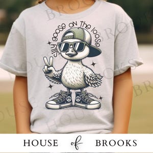 K&ouml;nnte beinhalten: Ein hellgraues T-Shirt mit einer Cartoon-Illustration einer Gans, die eine Sonnenbrille, eine Baseballkappe nach hinten und Turnschuhe tr&auml;gt. Die Gans h&auml;lt zwei Finger in einem Friedenszeichen hoch. Der Text "Silly goose on the loose" ist um die Gans herumgedruckt.
