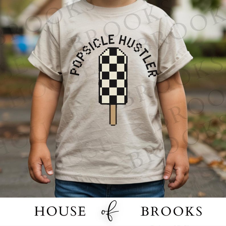 May include: A light tan t-shirt featuring the text "POPSICLE HUSTLER" arched above a pixelated black and white checkerboard popsicle graphic. The shirt has short sleeves and is worn by a child. The brand "HOUSE of BROOKS" is printed at the bottom.