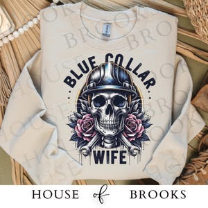 May include: A light beige sweatshirt with a skull wearing a hard hat and the text "Blue Collar Wife".