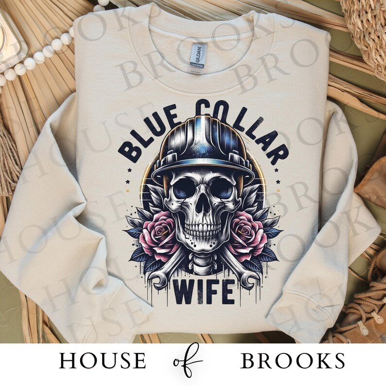May include: A light beige sweatshirt with a skull wearing a hard hat and the text "Blue Collar Wife".