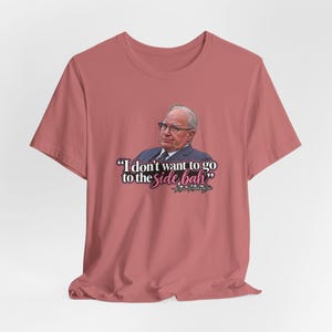 May include: A dusty rose crew neck t-shirt featuring a graphic of a man in a suit and glasses. Below the image, the text reads: "I don't want to go to the side bah" followed by "- Kevin Reddington" in a mix of black and pink script and print fonts.