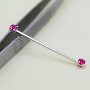Puede incluir: Una joya corporal de barra recta que presenta un eje de metal fino en tono plateado con una pequeña piedra preciosa redonda de color rosa vibrante engastada en cuatro garras en cada extremo, sujeta por pinzas de metal con patrón texturizado.