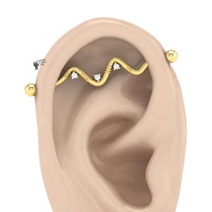 May include: A gold-colored industrial barbell earring with a wavy design and small, clear gemstones. The earring is shown on a rendered ear, with a gold ball on each end of the barbell.