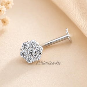 May include: A silver-colored lip piercing stud with a cluster of small, clear gemstones forming a flower shape. The stud is set against a soft, beige background with the text "Shirish Sparkle".