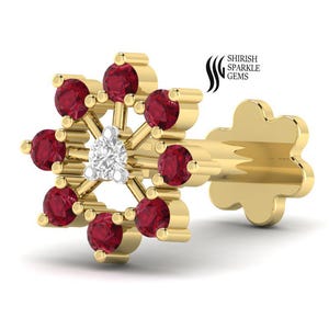 May include: A gold-colored nose stud with a floral design. The stud features a central white diamond surrounded by ten round, red gemstones. The post has a flower-shaped end. The brand name "SHRISH SPARKLE GEMS" is in the upper right corner.