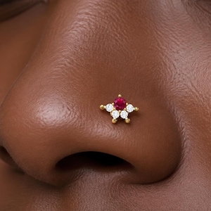 Può includere: Un piercing da naso in oro con una gemma rossa centrale a taglio rotondo, circondata da quattro pietre bianche trasparenti più piccole disposte a stella. Il gioiello è montato su una base in metallo dorato lucido posizionata sul naso.