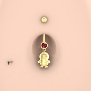 Op de afbeelding: Een gouden navelpiercing met een kleine, ronde, donkerroze edelsteen in een ronde zetting. Onder de edelsteen hangt een gepolijste gouden bedel in de vorm van een gestileerd kuikentje. Het sieraad wordt getoond op een gladde, lichte huid.