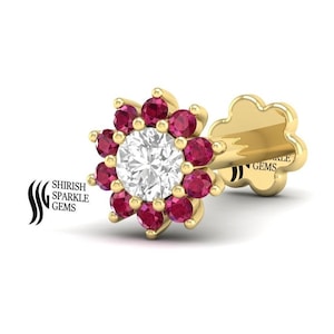 Pode incluir: Um anel de nariz de ouro com uma pedra branca redonda cercada por um círculo de pedras vermelhas. O anel de nariz tem a forma de uma flor.