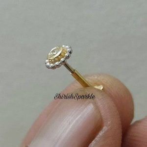 May include: A gold and silver nose stud with a small round clear stone in the center. The stud is surrounded by a beaded halo.