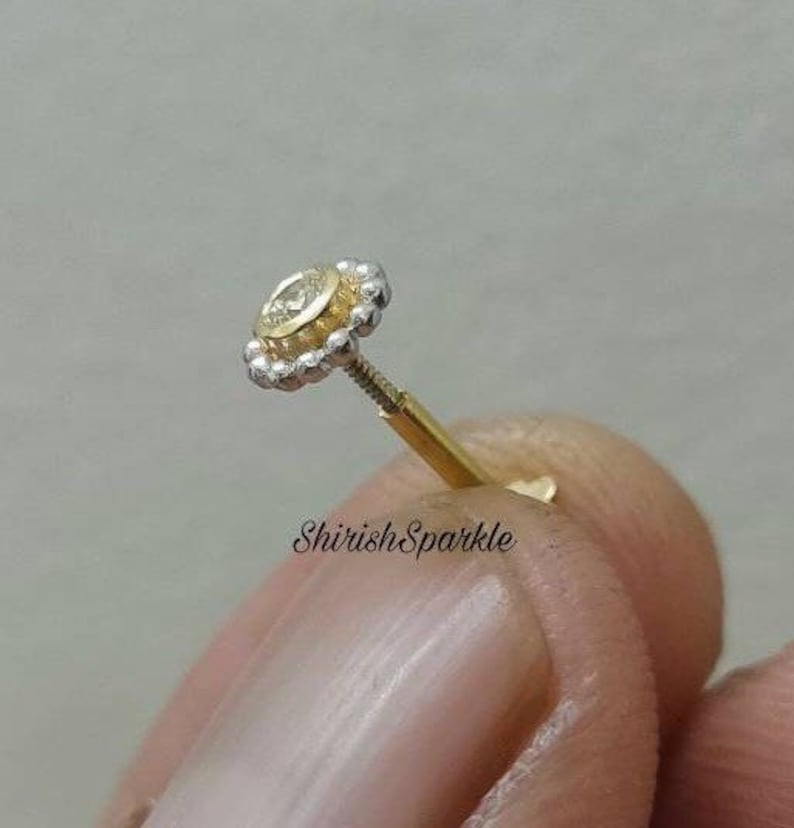 May include: A gold and silver nose stud with a small round clear stone in the center. The stud is surrounded by a beaded halo.