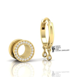 Puede incluir: Un par de pendientes de oro con un dije de diamante colgante. Los pendientes están hechos de oro y tienen un diseño circular con una fila de diamantes alrededor del borde. Los pendientes también están disponibles en estilo aro.