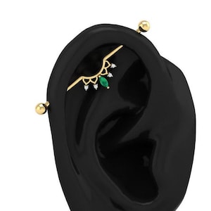 Puede incluir: Barbell de oro con una esmeralda verde y diamantes transparentes, usado como piercing de oreja.
