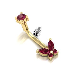 Può includere: Un piercing all'ombelico color oro con una barra curva e un gruppo di pietre preziose rosse. Il design include una grande gemma rotonda in alto e una disposizione floreale di gemme più piccole in basso. Il testo "SHIRISH SPARKLE GEMS" è visibile.