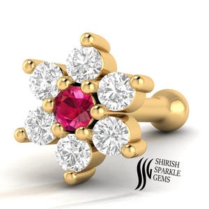 May include: A gold-colored flower-shaped stud earring with a central red gemstone surrounded by six white gemstones. The earring has a post and a round backing. The text "SHIRISH SPARKLE GEMS" is visible.
