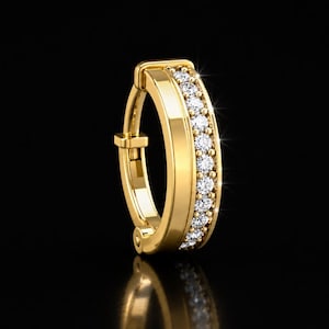 May include: A gold hoop earring featuring a hinged closure mechanism and a single row of small, clear, round-cut stones embedded along the outer edge. The polished metal surface reflects light, highlighting the circular design and the stones.
