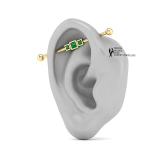 May include: Gold industrial barbell with three emerald green square gemstones. The barbell is worn in a person's ear.