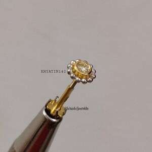 May include: A gold nose ring with a small, clear gemstone in the center. The ring is surrounded by a halo of small, white beads.