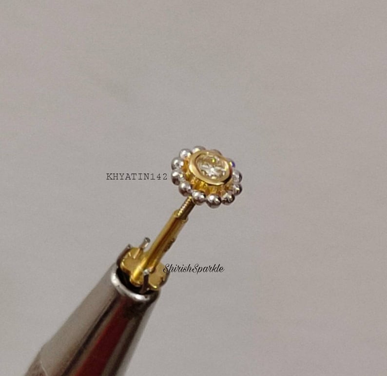 May include: A gold nose ring with a small, clear gemstone in the center. The ring is surrounded by a halo of small, white beads.