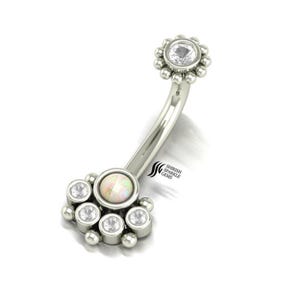 May include: A curved, silver-toned belly button ring with a cluster of clear, round gemstones and a central opal-like stone. The top features a flower-shaped design with a large, clear gemstone. The brand name "Shirish Sparkle Gems" is visible.