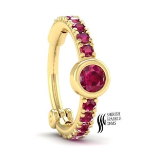 Pode incluir: Anel de nariz de ouro com uma grande pedra preciosa de rubi e pedras de rubi menores ao redor da banda. O nome da marca "Shirish Sparkle Gems" é visível no anel.