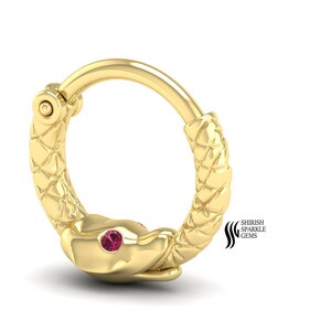 May include: A gold-colored hoop earring with a snake design. The snake's head features a small, red gemstone for an eye. The earring has a textured, scale-like pattern along its body. The jewelry is displayed on a white background.