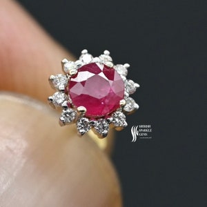 May include: A close-up of a ruby and diamond cluster ring. The ring features a large, round, deep red ruby center stone, surrounded by a halo of smaller, sparkling diamonds. The setting appears to be gold or a gold-toned metal.