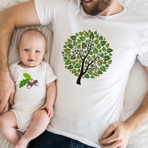 May include: A white t-shirt with a green tree design made of leaves. A baby is wearing a white onesie with a brown acorn design.