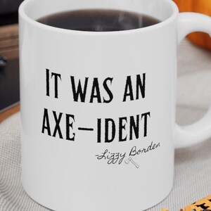 May include: White ceramic mug with the text "IT WAS AN AXE-IDENT" and "Lizzy Borden" in black. The mug is filled with a dark liquid and sits on a gray surface with Halloween-themed candies.