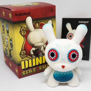Könnte beinhalten: Eine weiße Vinyl-Dunny-Figur mit blau gefärbten Ohren, großen roten und schwarzen Augen und einem blau-türkisfarbenen Brustdesign. Die Figur steht neben ihrer Box, die ein Zirkus-Thema und das Kidrobot-Logo aufweist.