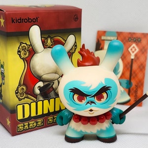 Könnte beinhalten: Eine weiße und blaue Dunny-Figur mit roten Haaren und einer roten und weißen Perlenkette. Die Figur hält einen Stab mit einem roten Kreuz oben. Die Figur steht auf einem weißen Hintergrund. Die Figur ist Teil der Kidrobot Dunny Side Show-Serie.